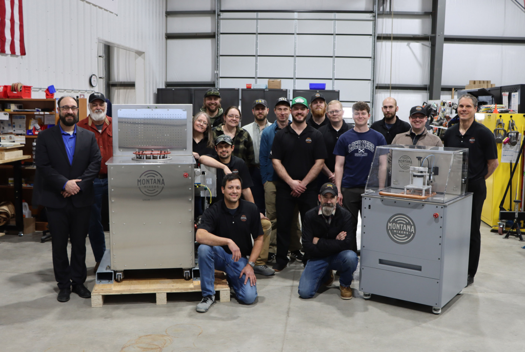 A group of people gather around two resonant mixing machines for a posed photograph in a manufacturing facility